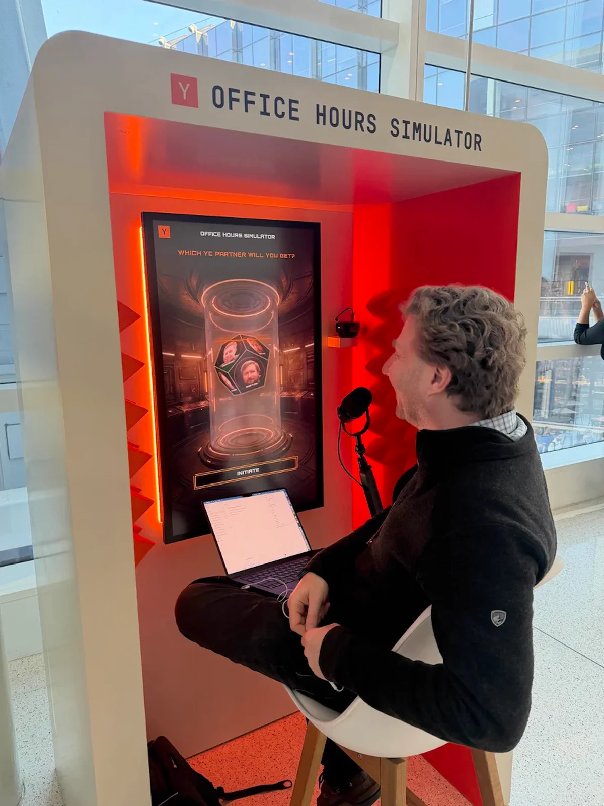 A user seated at YC’s orange Office Hour Simulator booth, with a microphone and a screen showing AI versions of YC partners.